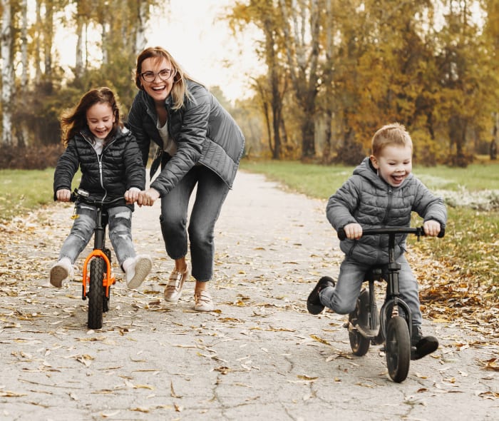A woman with two young children smiling and laughing as the kids ride their bikes along a tree lined path.