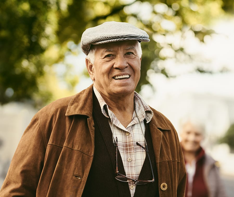 Older man with a hat on smiling for the camera. Enjoying a day out with a lady who is in the distance behind him.