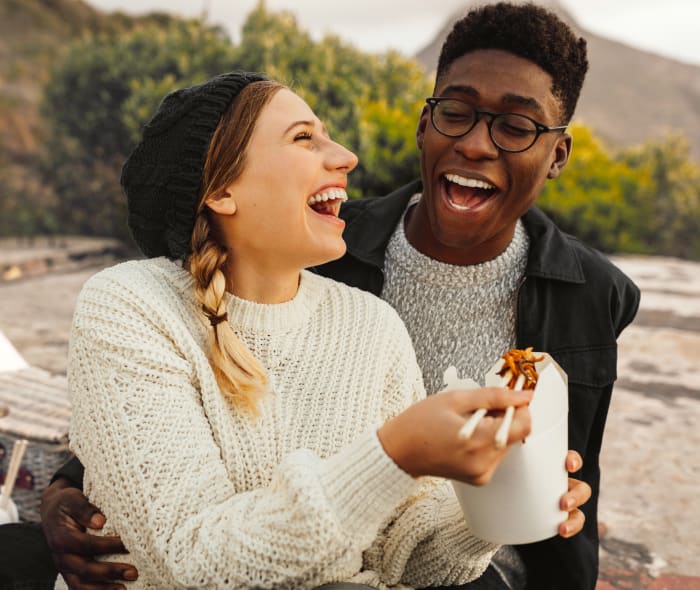 A young couple with healthy looking teeth laughing and sharing lunch on a beach.