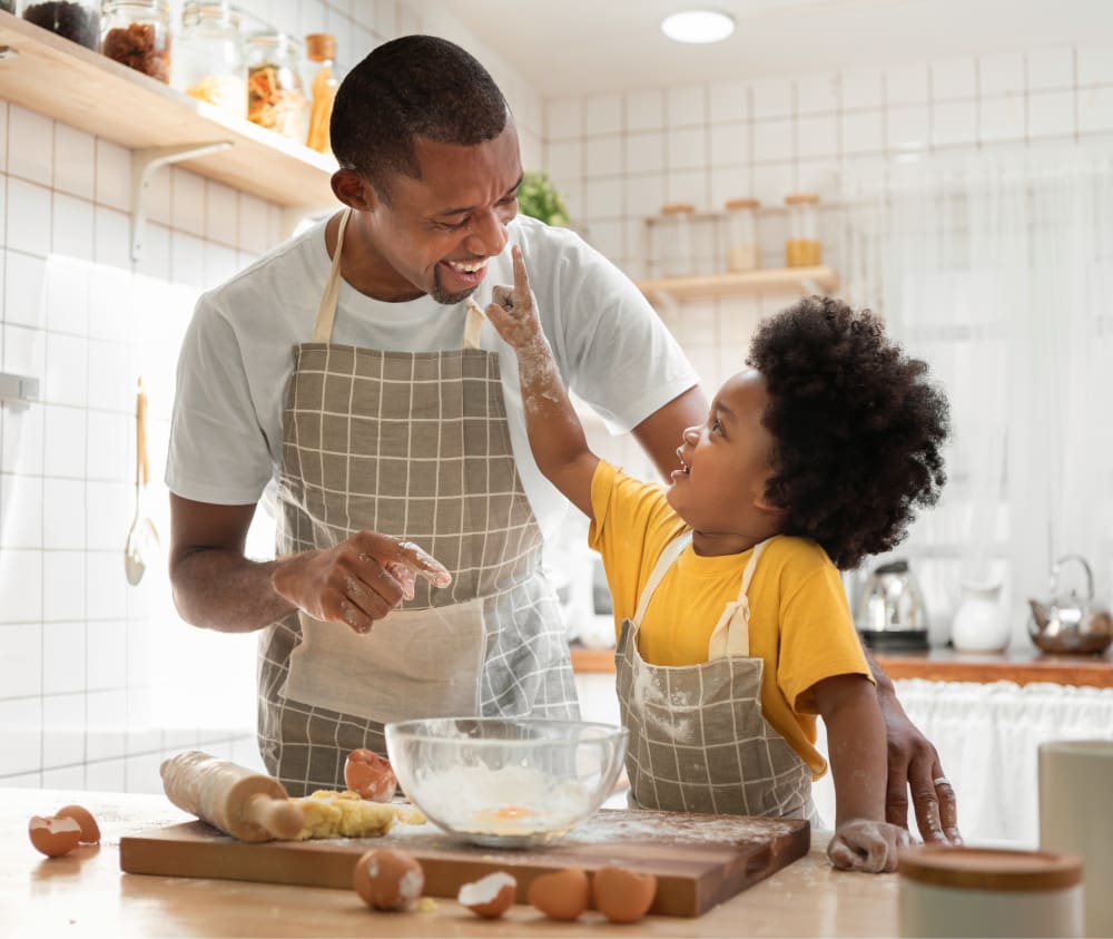 A father and young child with healthy looking teeth, smiling at each other as they are baking in the kitchen.