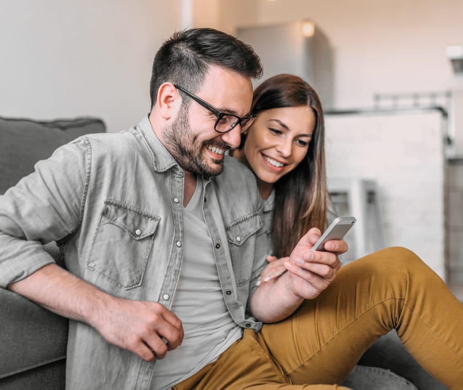 A couple with healthy looking teeth, sitting together on the living room floor smiling while looking at something on a phone.