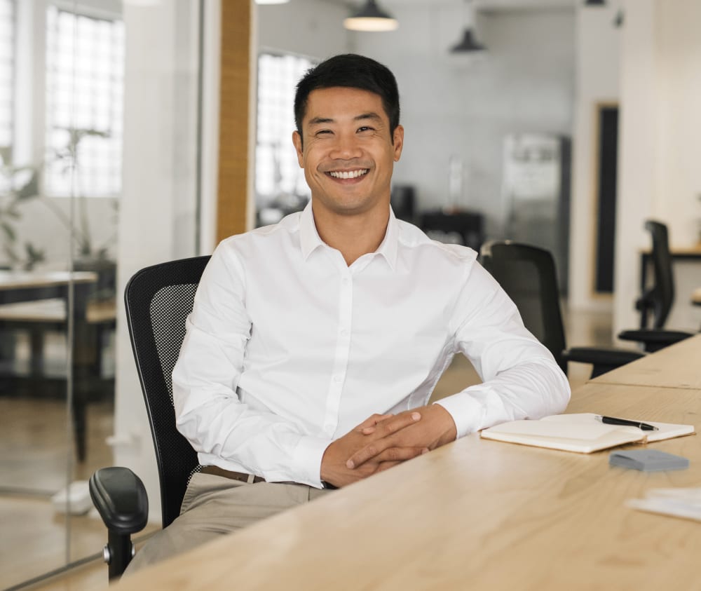 Man sitting at a desk in an office. He has clean, white teeth and is smiling happily at the camera.