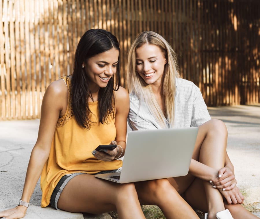 Two young ladies with beautiful smiles sitting together outside smiling and looking at a laptop.